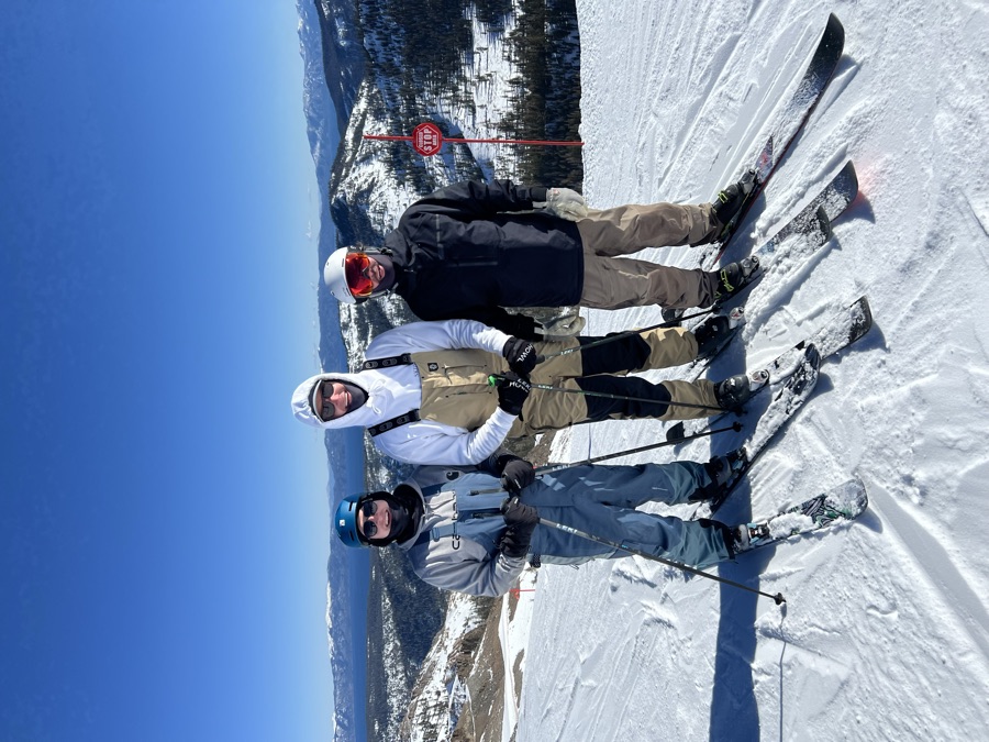 Three skiers at a resort summit beside a boundary sign, Lake Tahoe behind