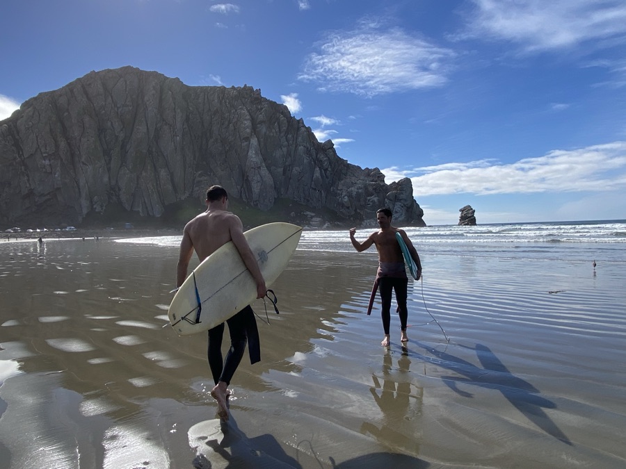 Two surfers carrying boards toward the water beneath Morro Rock on the California coast